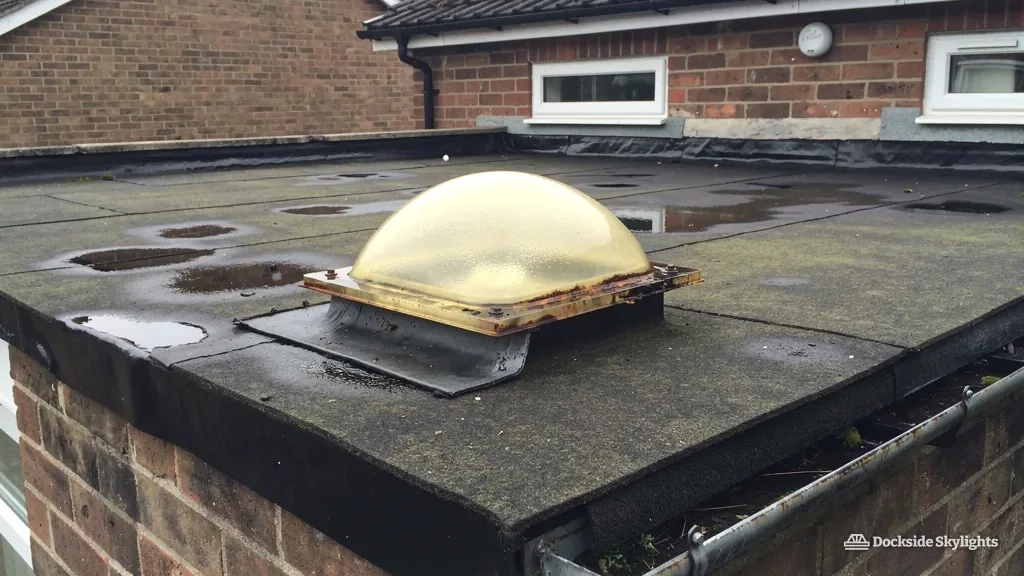 Yellowed plastic dome rooflight on a worn flat roof beside red-brick houses